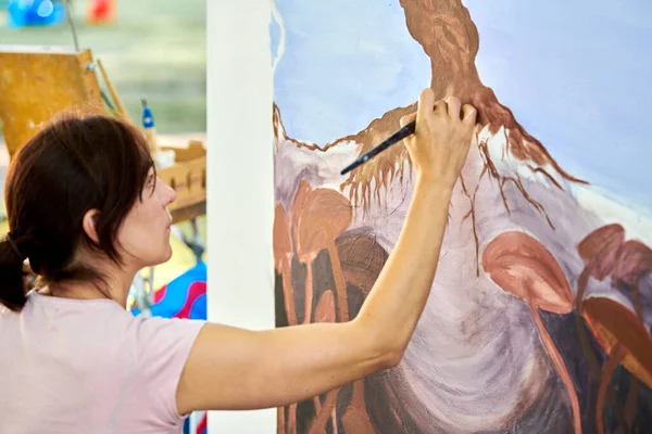 Girl artist draws with paint brush surreal nature landscape on white canvas at outdoor art painting festival, paintings art picture process, rear view. Woman artist paints atmospheric surreal picture