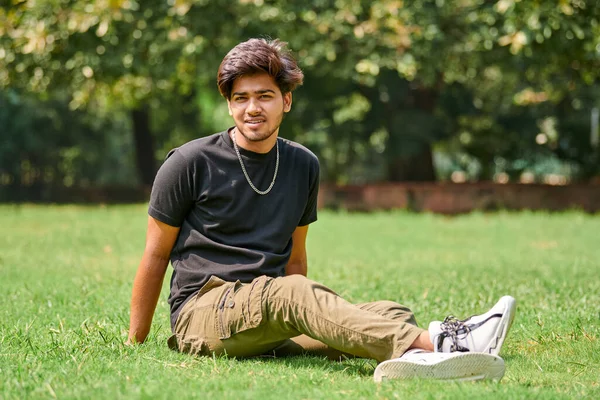 Smiling indian man candid portrait in black t shirt and silver neck chain sitting on green lawn in public park background, hindu attractive male portrait. Handsome indian man portrait with thick hair