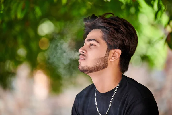 Attractive indian man smoker exhales cigarette smoke portrait in black t shirt and silver neck chain in public park, hindu male smoking close up portrait. Handsome indian man portrait with thick hair