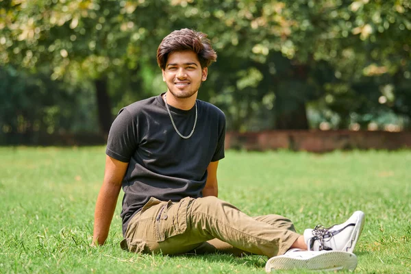 公園の背景に緑の芝生の上に座って黒のTシャツと銀の首のチェーンで笑顔インドの男率直な肖像画 魅力的な男性の肖像画をHindu 太い髪を持つハンサムなインド人の男性の肖像画 — ストック写真