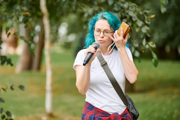 Young adult woman host of event in round glasses with turquoise dyed hair speaks into microphone at outdoor art exhibition, attractive female artist with bright appearance on green background