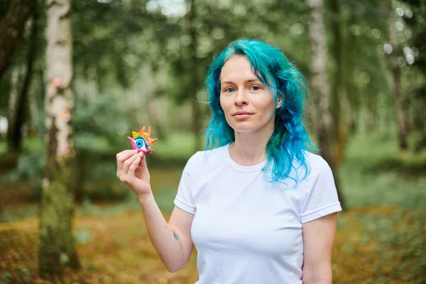 stock image Young woman with turquoise dyed hair in white T shirt holds handmade colorful eye amulet on green park background, art handmade artist on outdoor exhibition. Female handmade artist with eye art object