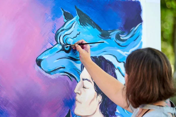 Woman artist hand holds paint brush and draws surreal fantasy image on white canvas at outdoor art painting festival, paintings art picture process. Woman artist paints atmospheric surreal picture