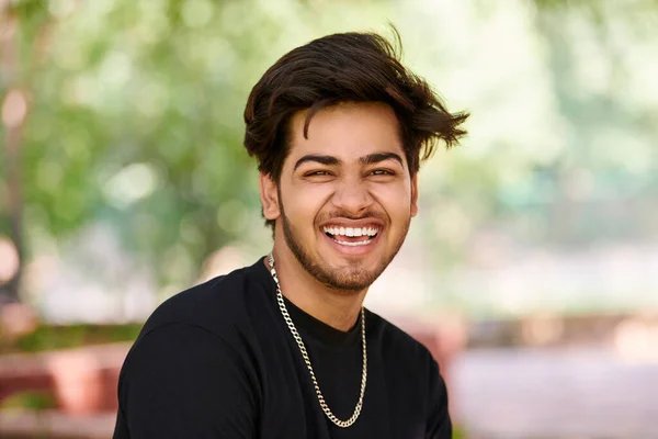 Smiling young indian man candid portrait in black t shirt and silver neck chain on outdoor public park background, close up hindu attractive male portrait. Handsome indian man portrait with thick hair