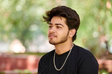 Siyah tişörtlü çekici genç Hintli adam portresi ve açık yeşil park arka planında gümüş boyun zinciri, Hindu erkek portresini kapat. Halk parkında gür saçlı yakışıklı Hintli adam portresi