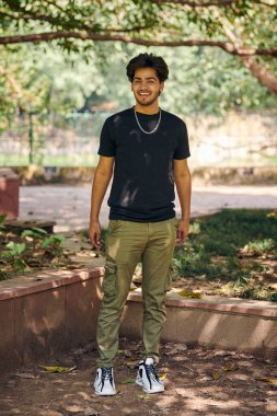 Çekici, gülümseyen Hintli adam. Siyah tişörtlü tam boy portre ve yeşil parktaki gümüş boyun zinciri, Hindu erkek portresi. Kamu parkında gür saçlı yakışıklı Hintli adam portresi.