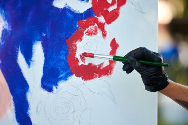 Siyah eldivenli sanatçı eli boya fırçasını tutuyor ve açık hava resim festivalinde beyaz tuval üzerine soyut gerçeküstü resimler çiziyor, resim resim süreci. Sanatçı atmosferik resim çizer