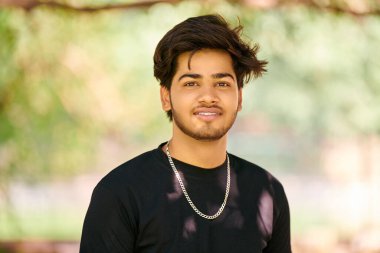 Siyah tişörtlü, gümüş boyunlu, halka açık park arka planında gülümseyen Hintli genç adam portresi, Hindu çekici erkek portresi. Kalın saçlı, yakışıklı Hintli adam portresi.