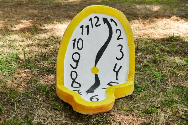 Melting clock art object lying on ground in forest at outdoor art exhibition, atmospheric surreal concept about fluidity of time in dream world. Bent clock with yellow edging and white clock face