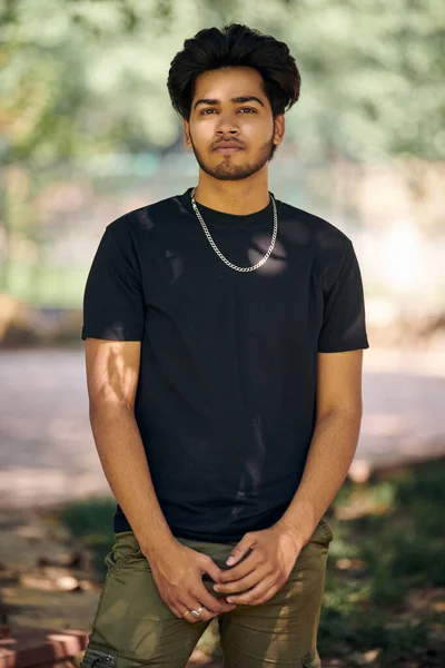 Attractive young indian man portrait in black t shirt and silver neck chain on outdoor green park background, close up hindu male portrait. Handsome indian man portrait with thick hair in public park