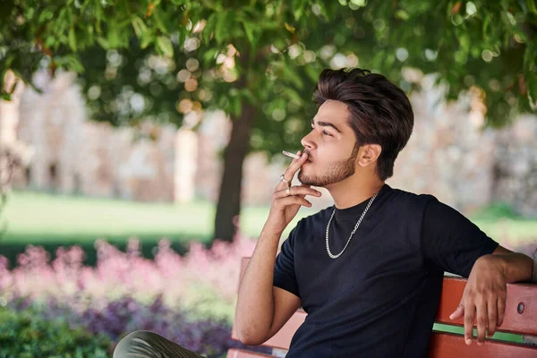 Young indian man smoker portrait in black t shirt and silver neck chain sitting on bench in public park, hindu male smoking portrait. Handsome indian man portrait with thick hair in city park