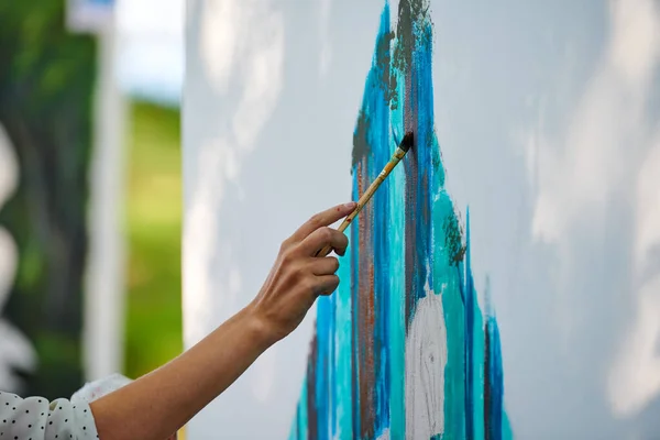 Artist hand holds paint brush and draws abstract surreal image on white canvas at outdoor art painting festival, paintings art picture process. Artist paints atmospheric picture