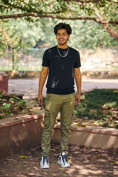 Attractive smiling indian man full height portrait in black t shirt and silver neck chain in green park, hindu male portrait. Handsome indian man outdoor portrait with thick hair in public park