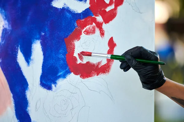 Siyah eldivenli sanatçı eli boya fırçasını tutuyor ve açık hava resim festivalinde beyaz tuval üzerine soyut gerçeküstü resimler çiziyor, resim resim süreci. Sanatçı atmosferik resim çizer