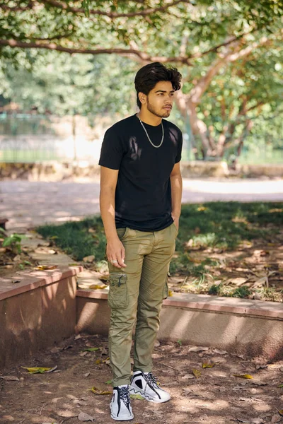 Attractive young indian man full height portrait in black t shirt and silver neck chain in green park, hindu male portrait. Handsome indian man outdoor portrait with thick hair in public park