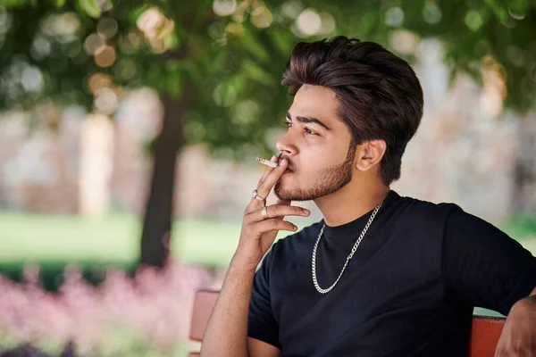 Young indian man smoker portrait in black t shirt and silver neck chain sitting on bench in public park, hindu male smoking close up portrait. Handsome indian man portrait with thick hair in city park