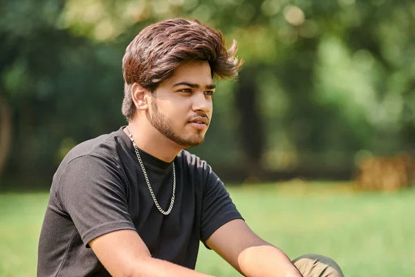 公共公園の緑の芝生の上に座って黒のTシャツと銀の首のチェーンで魅力的な若いインドの男性の肖像画 閉じるHindu男性の肖像画 公園で厚い髪のハンサムなインド人の男性の肖像画 — ストック写真