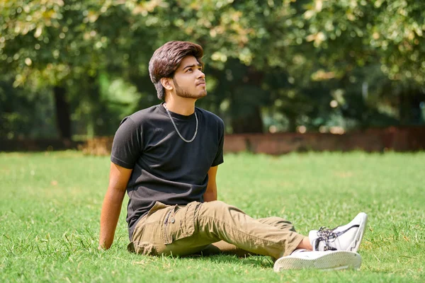 Attractive young indian man portrait in black t shirt and silver neck chain sitting on green lawn in public park, close up hindu male portrait. Handsome indian man portrait with thick hair in park