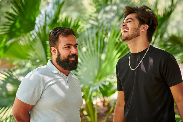 Два смешных индийских мужчины смеются и говорят на хинди в общественном парке зеленые пальмовые листья фона, настоящая мужская дружба. Два забавных индийских парня громко смеются на улице, гуляя в парке.