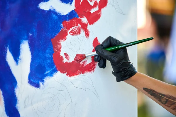 Artist hand in black gloves holds paint brush and draws abstract surreal image on white canvas at outdoor art painting festival, paintings art picture process. Artist paints atmospheric picture