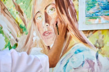 Ressam kız eli fırçayı tutuyor ve açık hava resim festivalinde beyaz tuvale gerçeküstü bir kadın portresi çiziyor, resim çiziyor. Kadın sanatçı atmosferik gerçeküstü resim çiziyor.