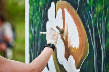 Ressam kız elinde boya fırçasıyla açık hava resim festivalinde beyaz tuvale yeşil bir manzara çiziyor, resim çiziyor. Kadın sanatçı atmosferik gerçeküstü resim çiziyor.