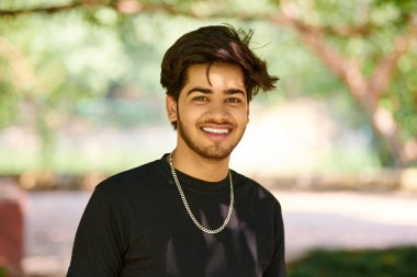 Siyah tişörtlü, gümüş boyunlu, halka açık park arka planında gülümseyen Hintli genç adam portresi, Hindu çekici erkek portresi. Kalın saçlı, yakışıklı Hintli adam portresi.