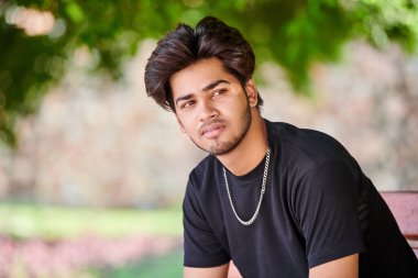 Çekici Hintli sigara içen adam siyah tişörtle sigara dumanı portresi ve halka açık parkta gümüş boyun zinciri veriyor. Hindu erkek, yakın portre sigara içiyor. Kalın saçlı, yakışıklı Hintli adam portresi.