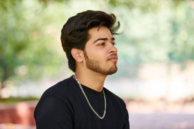 Siyah tişörtlü çekici genç Hintli adam portresi ve açık yeşil park arka planında gümüş boyun zinciri, Hindu erkek portresini kapat. Halk parkında gür saçlı yakışıklı Hintli adam portresi