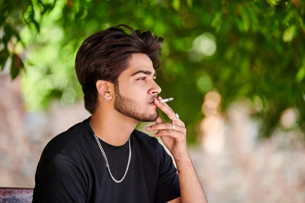 Young indian man smoker portrait in black t shirt and silver neck chain in public park, hindu male smoking close up portrait. Handsome indian man portrait with thick hair in city green park