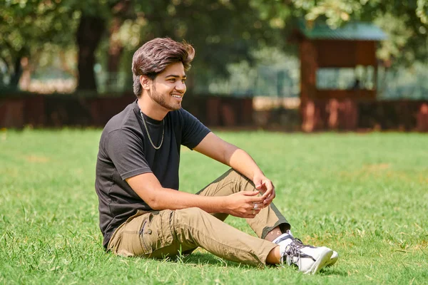 公園の背景に緑の芝生の上に座って黒のTシャツと銀の首のチェーンで笑顔インドの男率直な肖像画 魅力的な男性の肖像画をHindu 太い髪を持つハンサムなインド人の男性の肖像画 — ストック写真