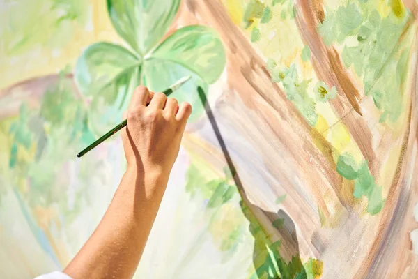 Girl artist hand holds paintbrush and draws green nature landscape on canvas at outdoor art painting festival, paintings art picture process. Woman artist paints atmospheric surreal picture