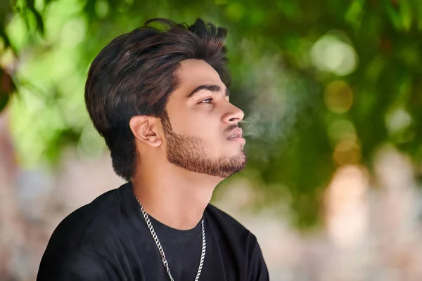 魅力的なインドの男性喫煙者は 公共の公園で黒いTシャツと銀の首のチェーンでタバコの煙の肖像画をExhales 密接な肖像画を吸うの背後にある男性 太い髪を持つハンサムなインド人の男性の肖像画 — ストック写真