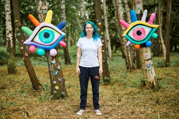Weird young adult woman with dyed turquoise hair in white T shirt near big eyes art installation in outdoor art exhibition in public park. Female handmade artist near colorful eyes art objects