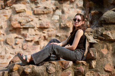 Çekici Doğu Asyalı siyah güneş gözlüklü ve siyah elbiseli kadın Kutub Minar kompleksindeki eski Hint mimari binalarının antik taş duvarında oturuyor, Hindistan 'daki samimi genç kız fotoğraf çekimi.