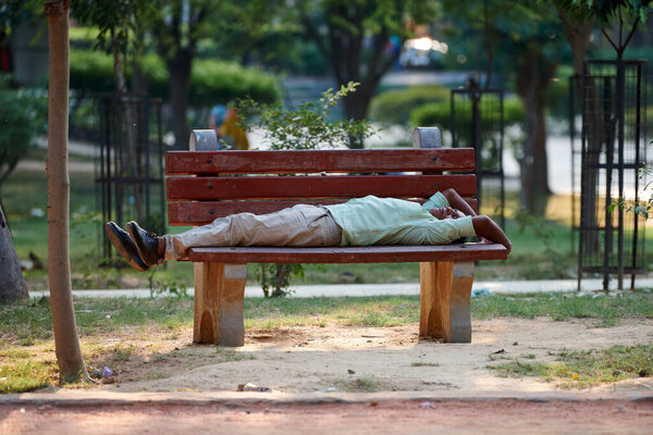 New Delhi, India - 11.11.2022 - Homeless indian man lies on bench in city park and resting, green trees background, houseless man sleeping in indian city park area, desperate disappointed homeless man