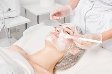 Güzellik uzmanı, güzellik salonlarında cilt bakımı için kadın yüzüne vitamin kremi maskesi, yaşlanma karşıtı kozmetik işlemler uyguluyor. Eldivenli kozmetik uzmanı maske takmak için kozmetik fırça tutuyor