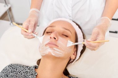 Kozmetik uzmanı, güzellik salonunda cildini yenilemek için kadın yüzüne kozmetik krem maskesi uyguluyor. Eldivenli güzellik uzmanı iki fırçalı kozmetik krem maskesi takıyor.