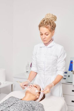 Kozmetik uzmanı, güzellik salonundaki kozmetik bakım ürünlerini nemlendirmek için kadın yüzüne estetik krem takıyor. Eldivenli güzellik uzmanı fırçayla kozmetik krem maskesi takıyor.