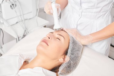 Cosmetologist makes ultrasound skin tightening for rejuvenation woman face using phonophoresis, anti aging cosmetic procedure with in beauty spa salon. Beautician makes ultrasonic skincare lifting clipart