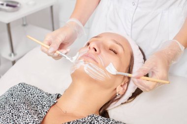 Kozmetik uzmanı, güzellik salonunda cildini yenilemek için kadın yüzüne kozmetik krem maskesi uyguluyor. Eldivenli güzellik uzmanı iki fırçalı kozmetik krem maskesi takıyor.