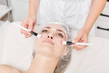 Güzellik uzmanı güzellik salonundaki cilt bakım kremi ve nemlendirici cilt bakımı için kadın derisine sürtünüyor. Kozmetik uzmanı elleri kadın yüzüne yüz kremi sürüyor.