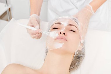 Güzellik uzmanı, güzellik salonlarında cilt bakımı için kadın yüzüne vitamin kremi maskesi, yaşlanma karşıtı kozmetik işlemler uyguluyor. Eldivenli kozmetik uzmanı maske takmak için kozmetik fırça tutuyor