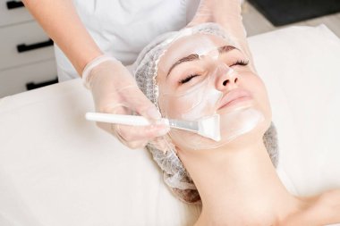 Güzellik uzmanı, güzellik salonlarında cilt bakımı için kadın yüzüne vitamin kremi maskesi, yaşlanma karşıtı kozmetik işlemler uyguluyor. Eldivenli kozmetik uzmanı maske takmak için kozmetik fırça tutuyor