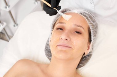 Güzellik uzmanı kadın yüzüne nemlendirici yüz derisi ve güzellik salonundaki yaşlanma karşıtı kozmetik prosedürleri için bal maskesi takıyor. Siyah eldivenli kozmetik uzmanı bal maskesi takmak için kozmetik fırça tutuyor.