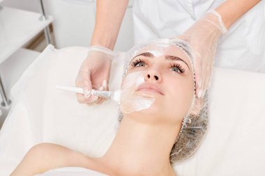 Güzellik uzmanı, güzellik salonundaki yaşlanma karşıtı kozmetik işlemler için cildini yenilemek için kadın yüzüne kozmetik krem maskesi sürüyor. Eldivenleri teslim eden kozmetik uzmanı maske takmak için kozmetik fırça tutuyor.