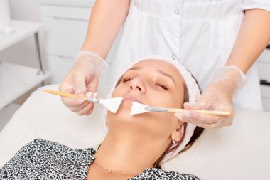 Güzellik uzmanı, güzellik salonunda estetik krem maskesini cilt tazeleme, nemlendirici kozmetik işlemleri için kadın yüzüne uyguluyor. Eldivenli kozmetik uzmanı iki fırçayla kozmetik krem maskesi takıyor.