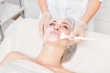 Güzellik uzmanı, güzellik salonundaki yaşlanma karşıtı kozmetik işlemler için cildini yenilemek için kadın yüzüne kozmetik krem maskesi sürüyor. Eldivenleri teslim eden kozmetik uzmanı maske takmak için kozmetik fırça tutuyor.
