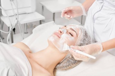 Güzellik uzmanı, güzellik salonundaki yaşlanma karşıtı kozmetik işlemler için cildini yenilemek için kadın yüzüne kozmetik krem maskesi sürüyor. Eldivenleri teslim eden kozmetik uzmanı maske takmak için kozmetik fırça tutuyor.