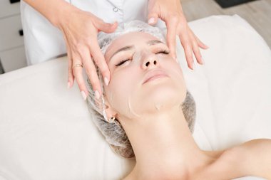 Güzellik uzmanı, güzellik salonundaki cilt bakımı için kadın yüzüne çarşaf maskesi, kırışıklık önleyici kozmetik prosedürler takıyor. Kozmetik uzmanı kadın yüzüne nemlendirici çarşaf maskesi uyguluyor.